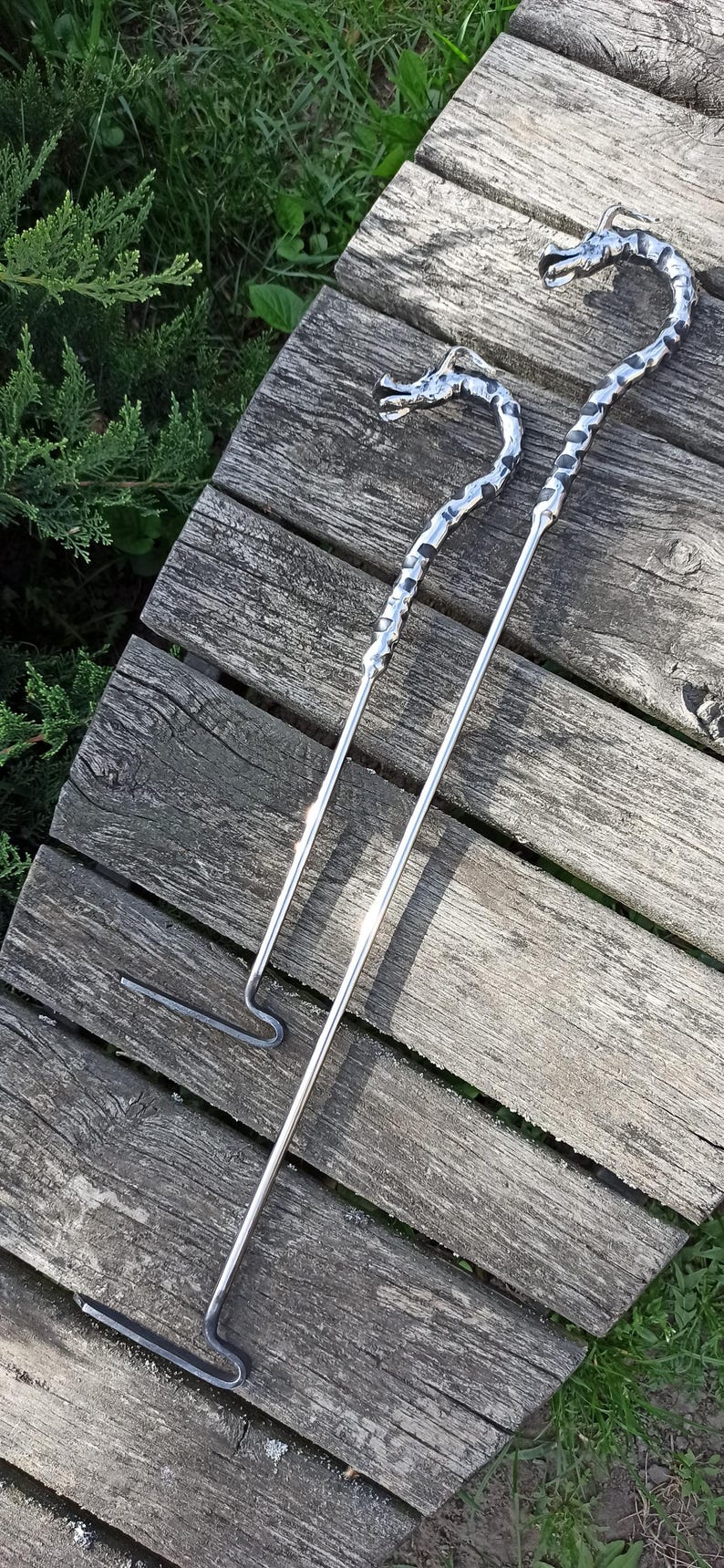 May include: Two metal fire pit tools with dragon head designs. One is a poker, and the other is a hook. The tools are silver and rest on weathered wooden planks.