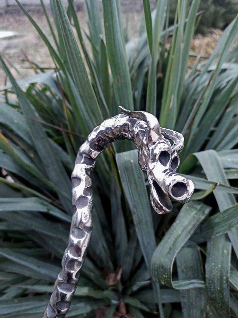 May include: A handcrafted, silver-colored metal dragon sculpture. The dragon's head is detailed with open nostrils and a curved horn. The textured body curves upwards, creating a unique decorative piece. The background features green foliage.