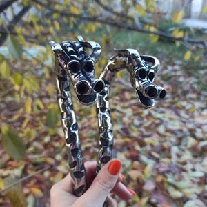 May include: A pair of silver-colored, dragon-shaped bracelets. The bracelets are held in a hand with red nail polish. The dragon heads have detailed features, including multiple eye and nostril openings. The background is blurred with fall foliage.