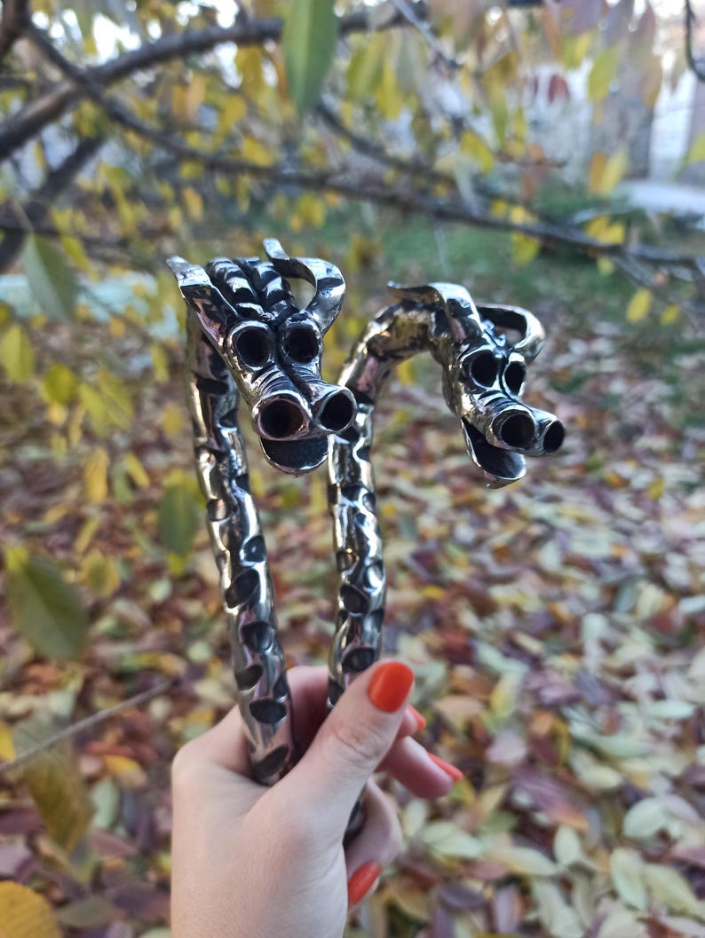 May include: A pair of silver-colored, dragon-shaped bracelets. The bracelets are held in a hand with red nail polish. The dragon heads have detailed features, including multiple eye and nostril openings. The background is blurred with fall foliage.