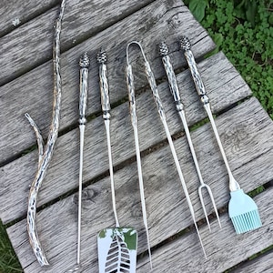 May include: A set of eight silver grilling tools with pine cone-shaped handles. The tools include a spatula, tongs, a fork, a brush, and three skewers. The tools are laid out on a wooden surface.