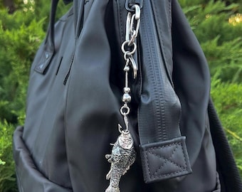 May include: A black backpack featuring a silver-toned metal keychain attached to the zipper pull. The keychain consists of a vertical chain with small spherical beads and a detailed silver fish charm suspended from a fishing hook.