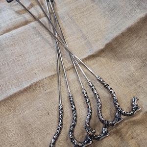 May include: A set of five metal fire pit tools. Each tool has a long, slender, silver shaft with a textured, curved handle and a black T-shaped top. The tools are arranged on a tan burlap surface.