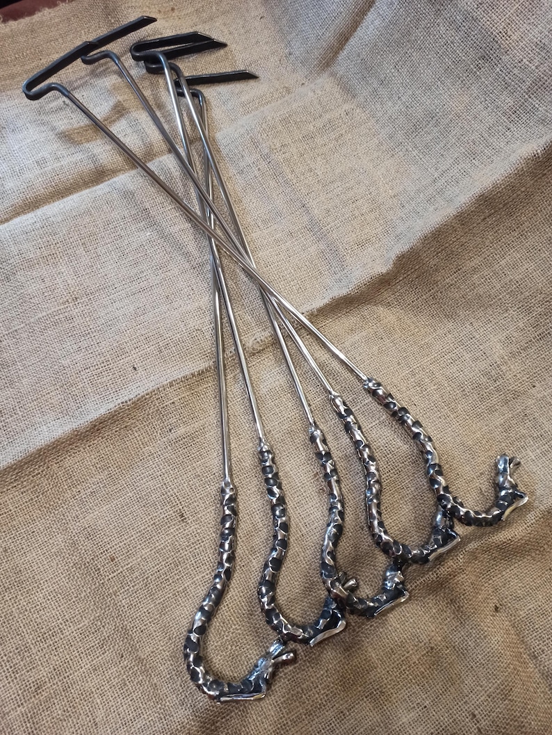 May include: A set of five metal fire pit tools. Each tool has a long, slender, silver shaft with a textured, curved handle and a black T-shaped top. The tools are arranged on a tan burlap surface.