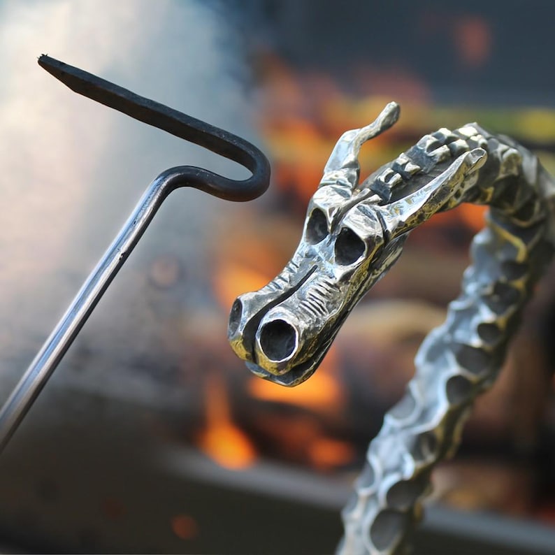 May include: Close-up of a metal fire poker and a decorative dragon head. The poker is black with a curved end. The dragon head is silver with detailed scales and eye sockets. The background is blurred with warm colors.