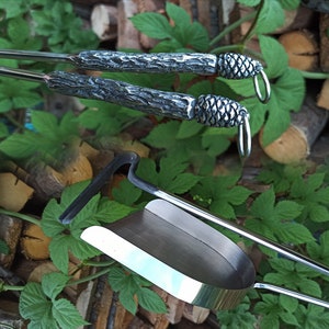 May include: A set of fireplace tools with a rustic design. The tools include a poker, tongs, and a shovel. The handles are made of metal and have a wood-like texture. The ends of the handles are shaped like pine cones.