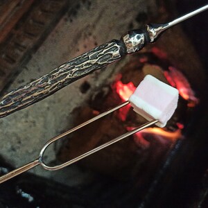 May include: A marshmallow on a metal roasting fork over a fire. The marshmallow is pink and white. A decorative metal roasting stick with a textured handle is in the background.