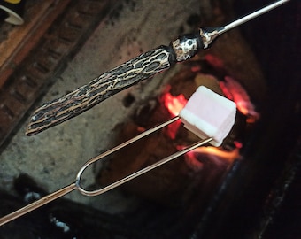 May include: A marshmallow roasting on a metal roasting fork over a fire. The marshmallow is pink and white. A decorative metal roasting stick with a textured handle is in the background.