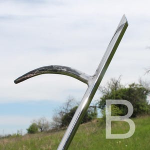 May include: A shiny, silver metal scythe blade with a curved, dark-tipped blade. The handle is angled and polished, reflecting the sky and surrounding greenery. The letter "B" is faintly visible in the background.
