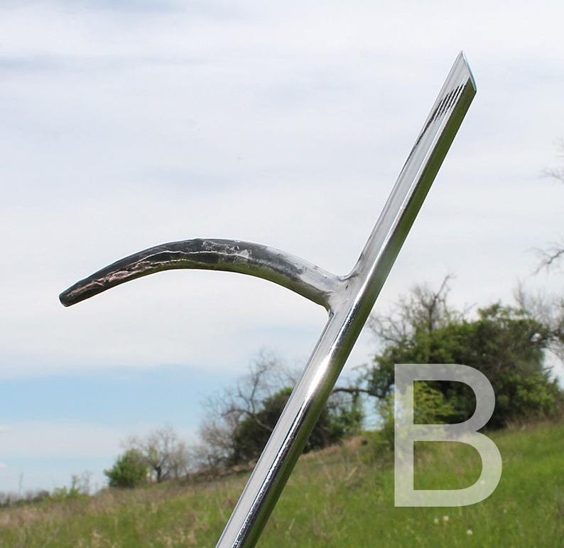 May include: A shiny, silver metal scythe blade with a curved, dark-tipped blade. The handle is angled and polished, reflecting the sky and surrounding greenery. The letter "B" is faintly visible in the background.