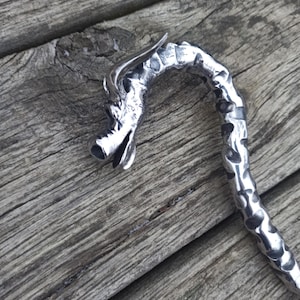 May include: A silver-colored dragon-shaped bookmark with a textured, curved body. The dragon's head is detailed with open mouth and horns. The bookmark is made of metal and rests on a weathered wooden surface.