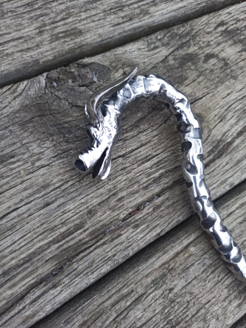 May include: A silver-colored dragon-shaped bookmark with a textured, curved body. The dragon's head is detailed with open mouth and horns. The bookmark is made of metal and rests on a weathered wooden surface.