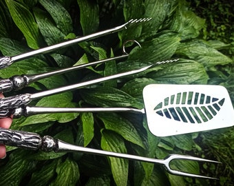 May include: A set of four stainless steel grilling tools with wooden handles. The tools include a spatula, tongs, a fork, and a basting brush. The spatula has a leaf-shaped cutout.