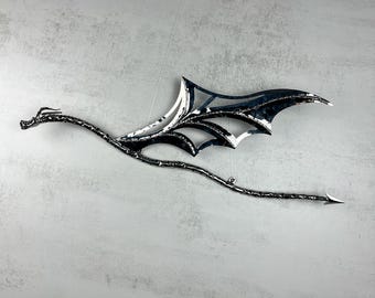 May include: A silver-toned metal wall hanging shaped like a dragon in flight. The dragon features a long, slender neck, a textured body, a pointed tail, and large, detailed wings with cut-out sections. The metal has a polished, reflective finish.