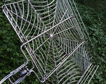 May include: A silver metal spider web shaped grilling basket with a handle. The handle is shaped like a spider.