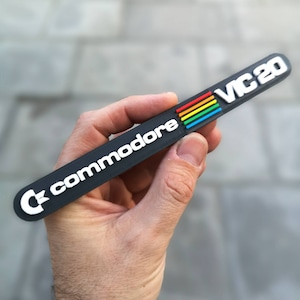 Puede incluir: Un llavero de plástico negro con el logotipo de Commodore y el texto "VC20" en letras blancas. El logotipo presenta un diseño de rayas arcoíris.