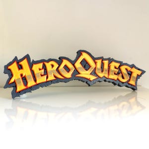 Może przedstawiać: Trójwymiarowa, wydrukowana tabliczka z napisem "HeroQuest" w stylizowanym kroju pisma. Litery są złote z czerwonym obramowaniem i umieszczone na czarnym tle.