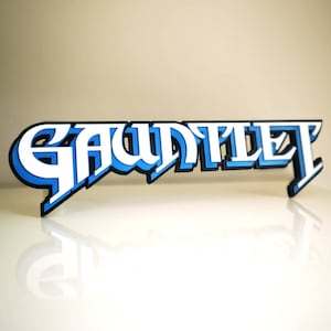 Könnte beinhalten: Ein 3D-gedrucktes Schild mit dem Wort "GAUNTLET" in weißen Buchstaben mit blauer Umrandung und schwarzem Schatten. Das Schild ist aus Kunststoff und hat eine glänzende Oberfläche.