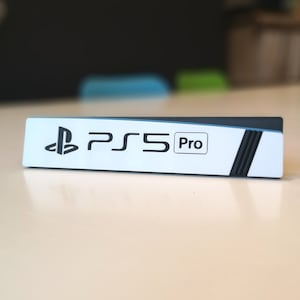 Puede incluir: Una consola rectangular blanca y negra con el logotipo de PlayStation y el texto "PS5 Pro" impreso en ella.