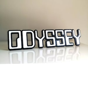 Könnte beinhalten: Das Wort "ODYSSEY" in 3D gedruckt in Weiß mit schwarzen Umrissen. Die Buchstaben sind erhaben und haben einen Retro-Futurismus-Stil.