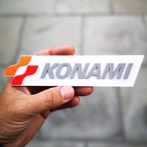 May include: A white rectangular sign with the word "KONAMI" in gray raised letters. A red and orange logo is on the left side of the sign.