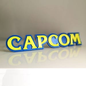 May include: A 3D printed logo of the word "CAPCOM" in blue and yellow. The letters are raised and have a textured surface.