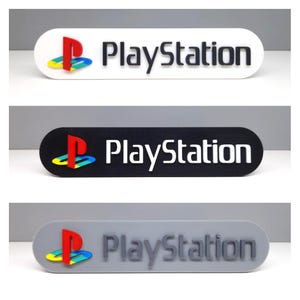 以下が含まれることがあります： 白、黒、グレーの3色のPlayStationロゴサイン。各サインには、赤、黄、青のPlayStationロゴと、黒またはグレーの「PlayStation」の文字が入っています。サインは角が丸い長方形です。