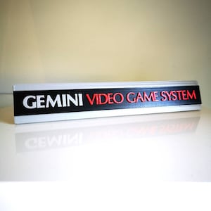 Könnte beinhalten: Ein silbernes und schwarzes Schild mit dem Text "GEMINI VIDEO GAME SYSTEM" in weißer und roter Schrift.