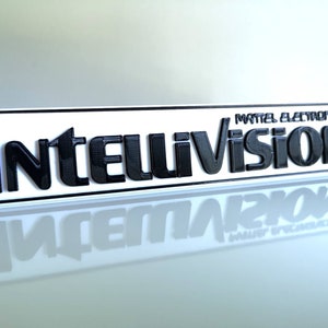 Könnte beinhalten: Ein rechteckiges Schild mit dem Wort "Intellivision" in fetten, schwarzen, erhabenen Buchstaben. Das Schild hat einen weißen Hintergrund und einen schwarzen Rand. Der Text "Mattel Electronics" steht über dem Haupttext.