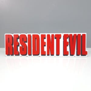 May include: A rectangular sign with the words "RESIDENT EVIL" in bold, red letters. The letters have a black outline and are set against a white background. The sign is on a white surface.