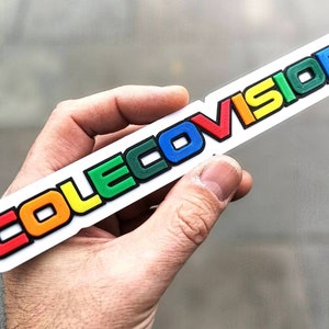 Könnte beinhalten: Ein weißer Aufkleber aus Kunststoff mit dem Wort "COLECOvision" in Regenbogenfarben. Die Buchstaben sind erhaben und haben einen schwarzen Umriss.