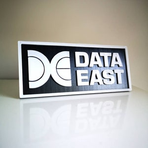 Könnte beinhalten: Ein schwarz-weißes Schild mit dem Text "DATA EAST" und einem stilisierten Logo aus einem ineinander verschlungenen "D" und "E".