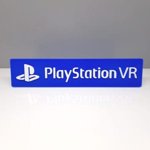 Puede incluir: Un letrero rectangular azul con el logotipo de PlayStation y la inscripción "PlayStation VR" en blanco. El letrero está sobre una superficie blanca con un fondo gris.