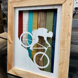 K&ouml;nnte beinhalten: Ein gerahmtes Wandbild aus Holz mit einer wei&szlig;en Silhouette einer Person, die mit dem Fahrrad einen H&uuml;gel hinunterf&auml;hrt. Der Hintergrund besteht aus bunten Holzstreifen in den Farben Rot, Wei&szlig;, Blau, Gelb, Gr&uuml;n und Braun.