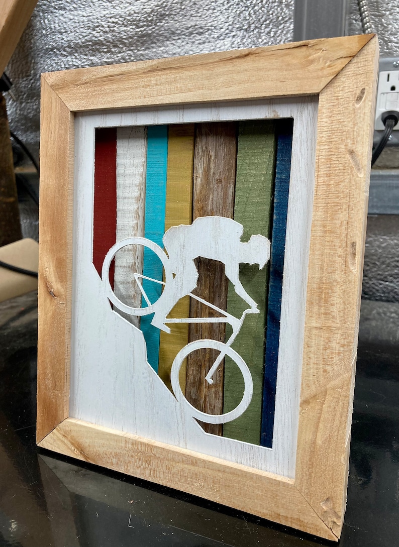 K&ouml;nnte beinhalten: Ein gerahmtes Wandbild aus Holz mit einer wei&szlig;en Silhouette einer Person, die mit dem Fahrrad einen H&uuml;gel hinunterf&auml;hrt. Der Hintergrund besteht aus bunten Holzstreifen in den Farben Rot, Wei&szlig;, Blau, Gelb, Gr&uuml;n und Braun.