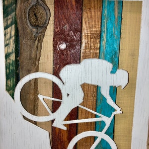 K&ouml;nnte beinhalten: Eine wei&szlig;e Silhouette einer Person, die auf einem Mountainbike auf einem Holzgrund f&auml;hrt. Das Holz ist in verschiedenen Braunt&ouml;nen, Blau- und Gr&uuml;nt&ouml;nen bemalt. Die Silhouette ist aus dem Holz geschnitten, wodurch ein 3D-Effekt entsteht.
