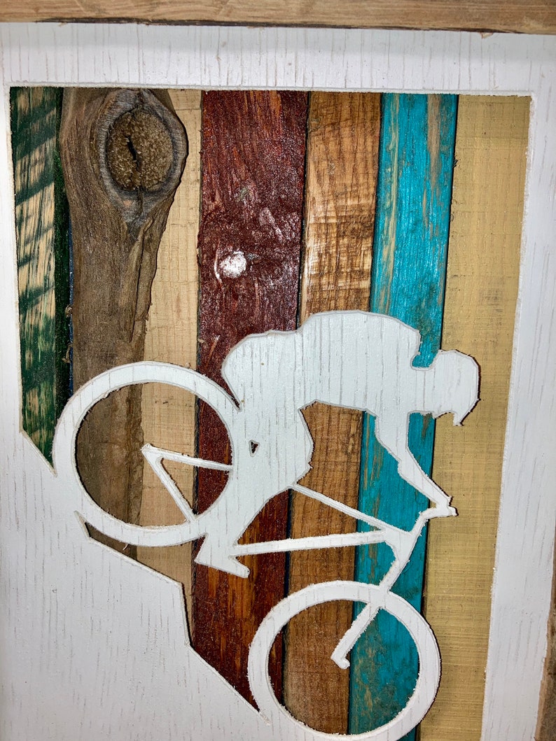 K&ouml;nnte beinhalten: Eine wei&szlig;e Silhouette einer Person, die auf einem Mountainbike auf einem Holzgrund f&auml;hrt. Das Holz ist in verschiedenen Braunt&ouml;nen, Blau- und Gr&uuml;nt&ouml;nen bemalt. Die Silhouette ist aus dem Holz geschnitten, wodurch ein 3D-Effekt entsteht.