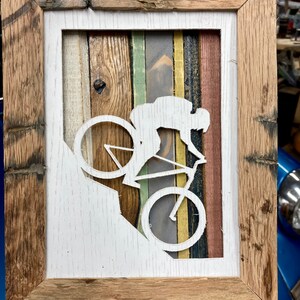K&ouml;nnte beinhalten: Eine wei&szlig;e Silhouette einer Person, die Fahrrad f&auml;hrt, ist aus einem Holzrahmen ausgeschnitten. Der Rahmen besteht aus wiederverwendetem Holz mit unterschiedlich farbigen Holzstreifen. Der Hintergrund ist wei&szlig;.