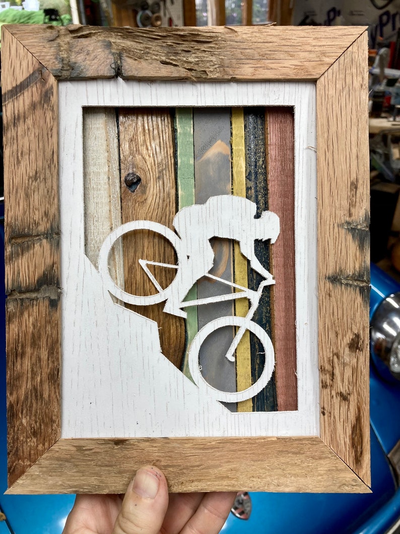 K&ouml;nnte beinhalten: Eine wei&szlig;e Silhouette einer Person, die Fahrrad f&auml;hrt, ist aus einem Holzrahmen ausgeschnitten. Der Rahmen besteht aus wiederverwendetem Holz mit unterschiedlich farbigen Holzstreifen. Der Hintergrund ist wei&szlig;.