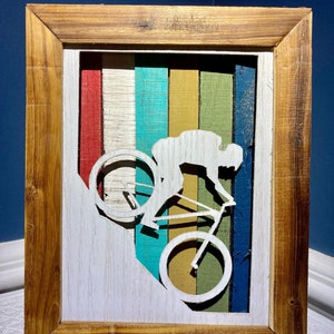 K&ouml;nnte beinhalten: Ein Wandbild in einem Holzrahmen mit der Silhouette einer Person, die einen H&uuml;gel mit dem Fahrrad hinunterf&auml;hrt. Der Hintergrund ist ein farbenfrohes Streifenmuster in Rot, Wei&szlig;, Blau, Gr&uuml;n und Gelb.