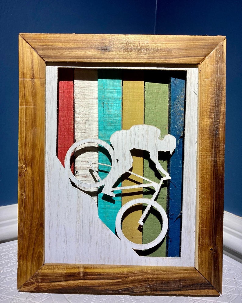 K&ouml;nnte beinhalten: Ein Wandbild in einem Holzrahmen mit der Silhouette einer Person, die einen H&uuml;gel mit dem Fahrrad hinunterf&auml;hrt. Der Hintergrund ist ein farbenfrohes Streifenmuster in Rot, Wei&szlig;, Blau, Gr&uuml;n und Gelb.