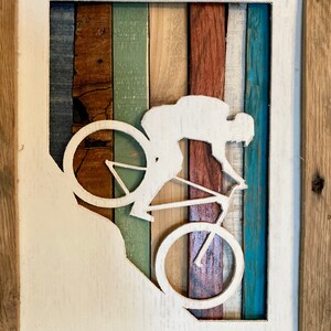 K&ouml;nnte beinhalten: Eine wei&szlig;e Silhouette einer Person, die Fahrrad f&auml;hrt, ist aus einem Holzrahmen ausgeschnitten. Der Rahmen besteht aus verschiedenen farbigen Holzstreifen, die ein gestreiftes Muster bilden. Der Hintergrund ist wei&szlig;.