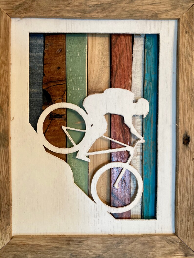 K&ouml;nnte beinhalten: Eine wei&szlig;e Silhouette einer Person, die Fahrrad f&auml;hrt, ist aus einem Holzrahmen ausgeschnitten. Der Rahmen besteht aus verschiedenen farbigen Holzstreifen, die ein gestreiftes Muster bilden. Der Hintergrund ist wei&szlig;.