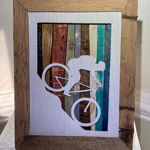 K&ouml;nnte beinhalten: Ein gerahmtes Holzbild mit einer wei&szlig;en Silhouette einer Person, die Fahrrad f&auml;hrt. Der Hintergrund besteht aus bunten Holzstreifen.