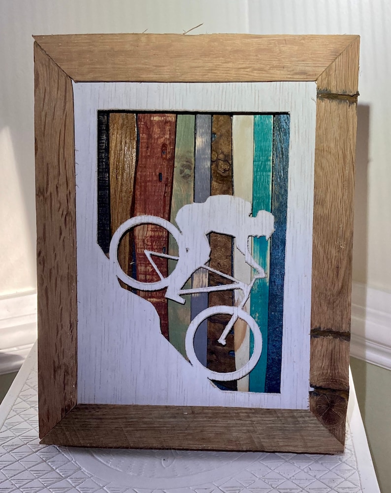 K&ouml;nnte beinhalten: Ein gerahmtes Holzbild mit einer wei&szlig;en Silhouette einer Person, die Fahrrad f&auml;hrt. Der Hintergrund besteht aus bunten Holzstreifen.