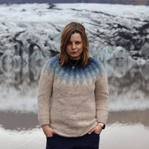 Könnte beinhalten: Eine Frau in einem beigefarbenen Pullover mit einem blauen und weißen geometrischen Muster steht vor einem Gletscher. Sie trägt eine silberne Halskette und hat die Hände in den Taschen.