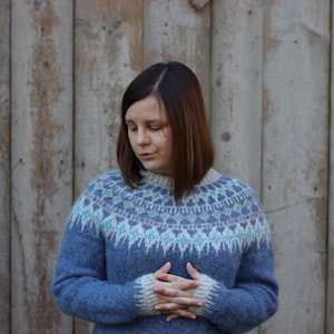 Puede incluir: Una mujer con un suéter azul y blanco con un diseño geométrico. El suéter tiene un cuello alto y mangas largas.