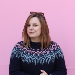 Può includere: Una donna indossa un maglione blu scuro con un motivo geometrico rosa e blu. Il maglione ha un collo alto e maniche lunghe. La donna è in piedi davanti a un muro rosa.