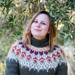 Può includere: Una donna indossa un maglione verde, marrone, bianco e rosso con collo alto. È in piedi davanti a uno sfondo verde di foglie.