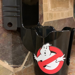 May include: Black plastic cup holder with a Ghostbusters logo featuring a white ghost with a red no symbol. The holder is mounted to a wall.